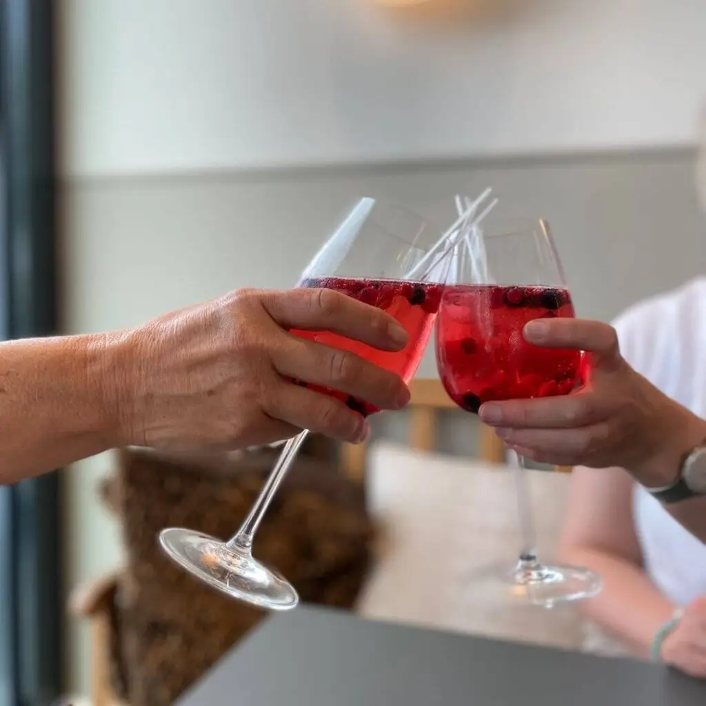 Zwei Hände stoßen mit rosaroten Beeren-Cocktails im Weinglas an – erfrischender Sommer-Aperitif mit Beeren und Strohhalm