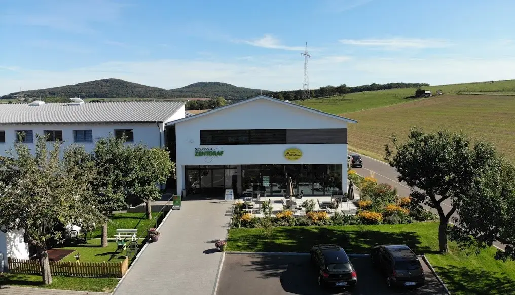 Außenansicht Schuhhaus Zentgraf mit Terrasse, Garten und Parkplätzen vor ländlicher Kulisse mit Hügeln