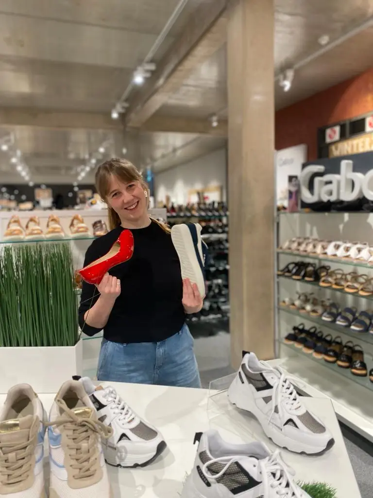 Frau im Schuhgeschäft hält roten High Heel und weißen Sneaker, Schuhregale und Display mit Sneakern im Hintergrund