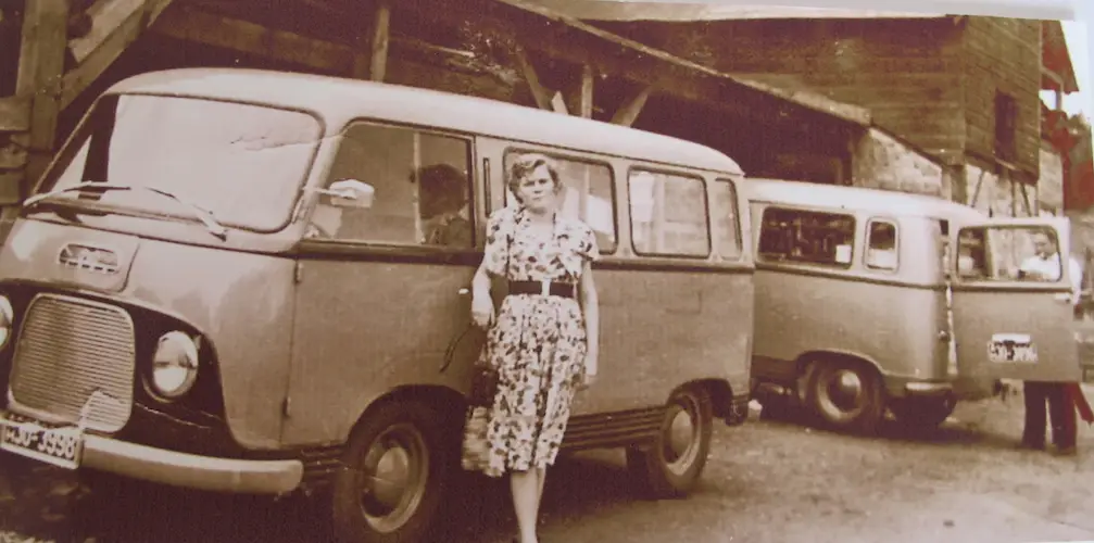 Sepia-Foto: Frau im Blumenkleid steht neben zwei alten Lieferwagen/Kleinbussen auf einem Hof