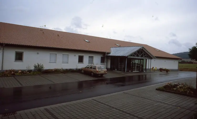 Eingang eines einstöckigen Bürogebäudes mit rotem Satteldach, Glasvorbau, nassem Parkplatz und parkendem Auto, ländlich