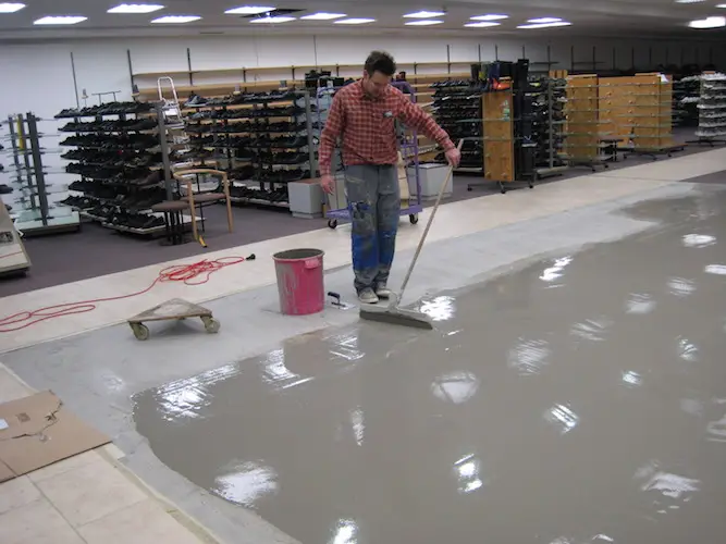 Handwerker trägt Fließestrich auf Ladenboden auf, glättet Oberfläche mit Rakel; Regale im Hintergrund