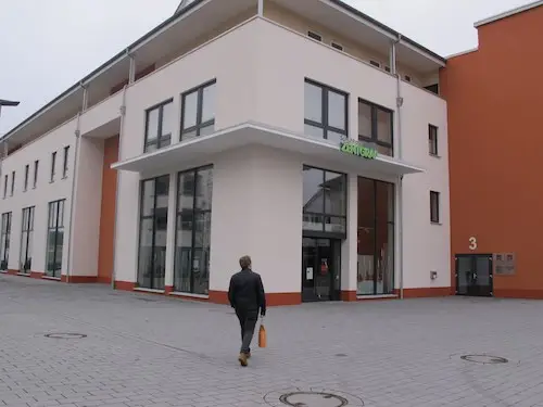 Person geht zum modernen Geschäftshaus mit Glasfront und Eingangsbereich auf gepflastertem Vorplatz